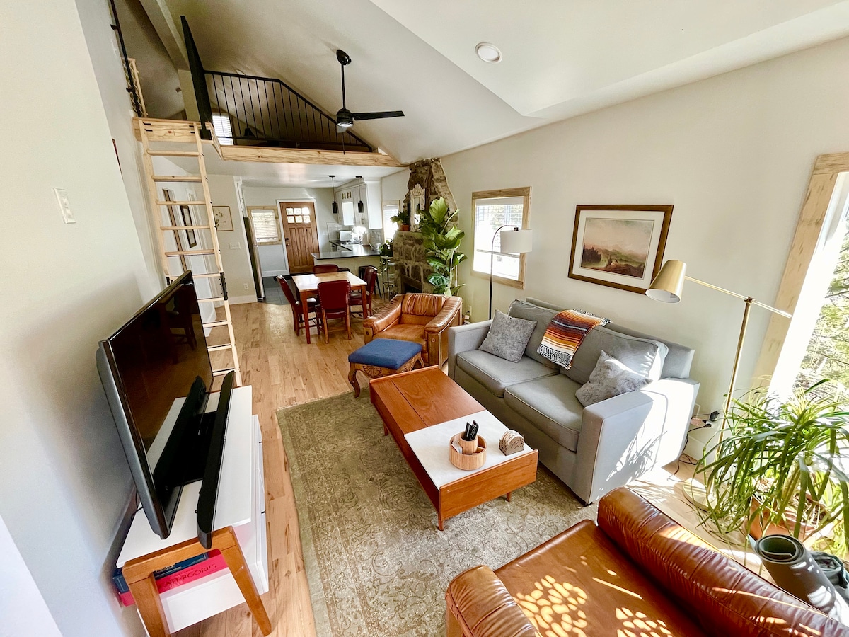 The living area showcases a comfortable two-person couch accompanied by a leather chair and warm wood accents. Natural light floods the space, highlighting the open-plan design that connects to the kitchen. A ladder provides access to the loft area, enhancing the cozy atmosphere.