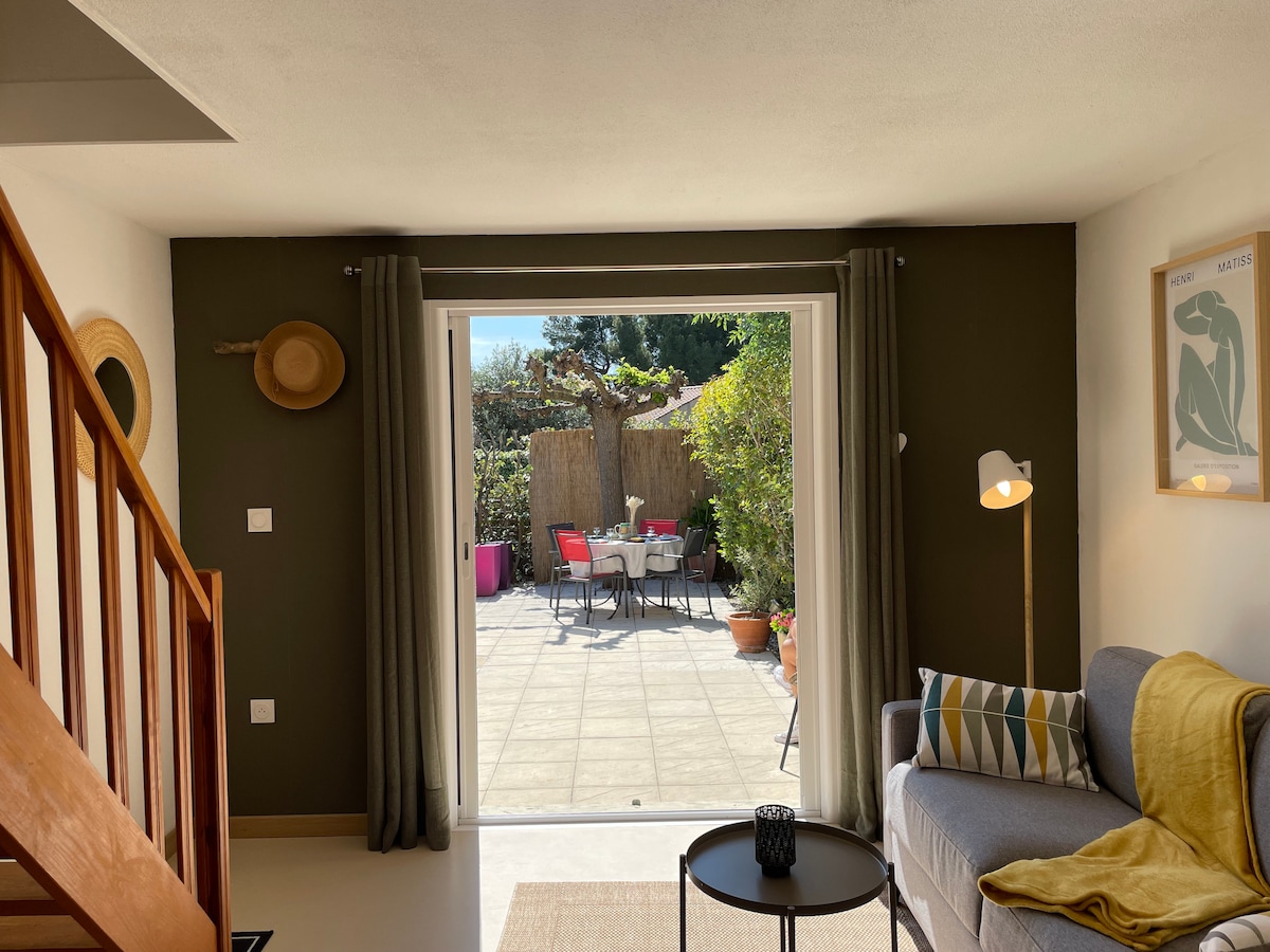 A bright living area is depicted, featuring a cozy sofa with patterned cushions. Large sliding doors open to a private outdoor patio, furnished with a dining table and chairs. Decor elements include a wall art piece and a stylish floor lamp, enhancing the modern ambiance.