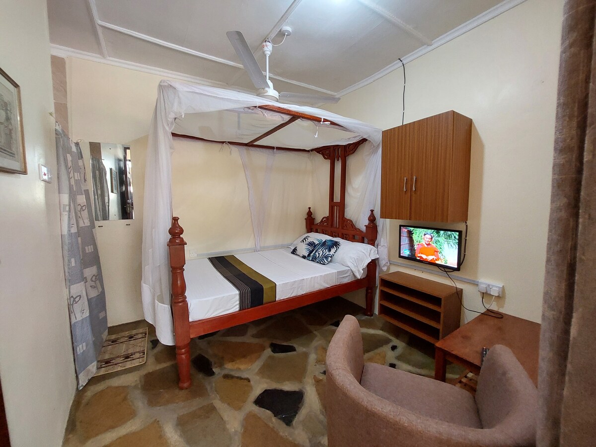A studio apartment features a four-poster bed with a canopy and a decorative throw, positioned alongside a wooden desk and a chair. A television is mounted on the wall, and the flooring consists of natural stone tiles. Shelving and closet space are also visible.