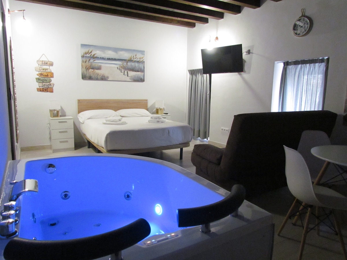A spacious room is highlighted by a private jacuzzi featuring illuminated blue water. A large bed, flanked by bedside tables, sits adjacent to a sofa. A dining table with four chairs is positioned nearby. Natural light enters through a window with gray curtains.