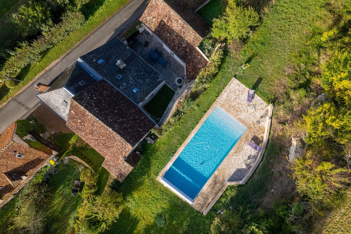 An aerial view captures the spacious property with a modern swimming pool adjacent to the house. The pool area is outlined with sun loungers, surrounded by manicured greenery and a pathway leading to the entrance of the residence.