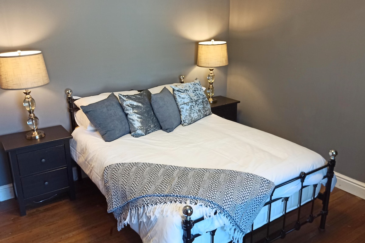A comfortable bedroom features a spacious bed with a white comforter and decorative pillows. Two bedside tables with lamps provide soft lighting. The walls are painted in a muted gray, enhancing the serene atmosphere. Wooden flooring complements the overall design.