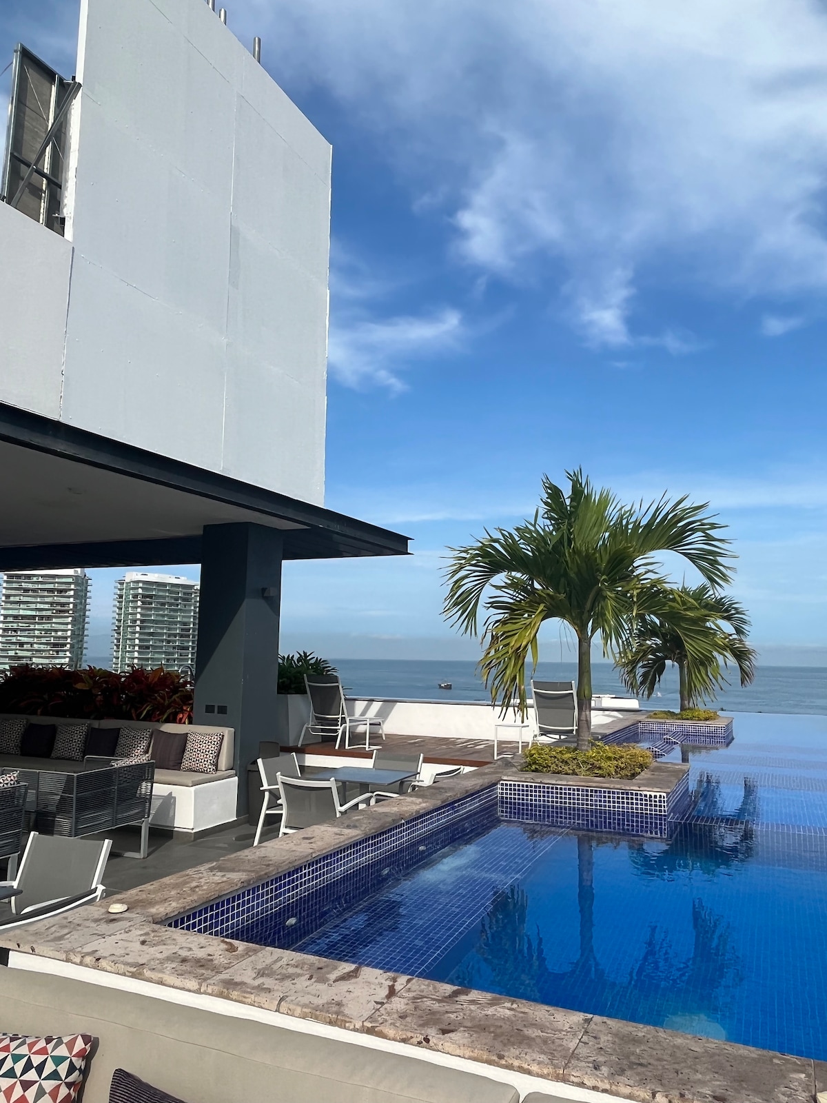An inviting rooftop space is highlighted by a shimmering pool that seamlessly blends with the horizon. Comfortable lounge chairs are arranged around the pool area, while lush greenery adds a touch of nature. A clear blue sky enhances the overall tranquil atmosphere.