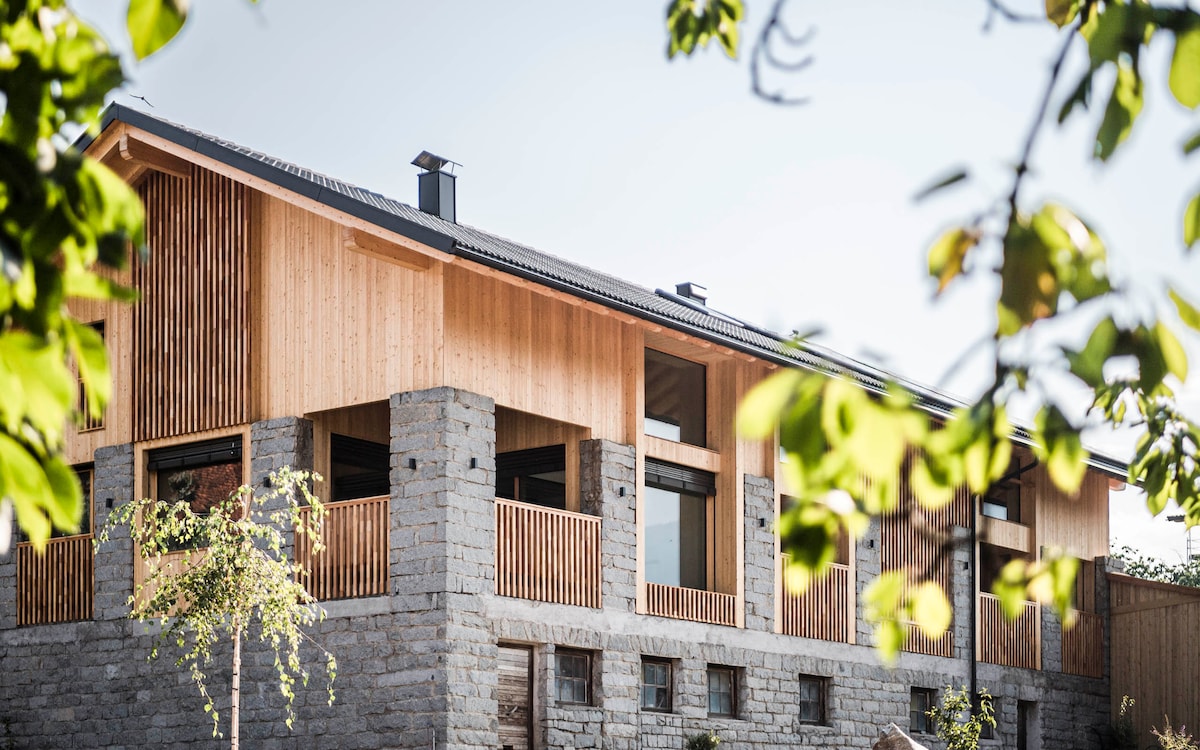 The chalet features a striking combination of wooden and stone elements. Large windows allow natural light to fill the interior, while a spacious balcony offers scenic views. Surrounding greenery frames the exterior, enhancing the tranquil atmosphere.