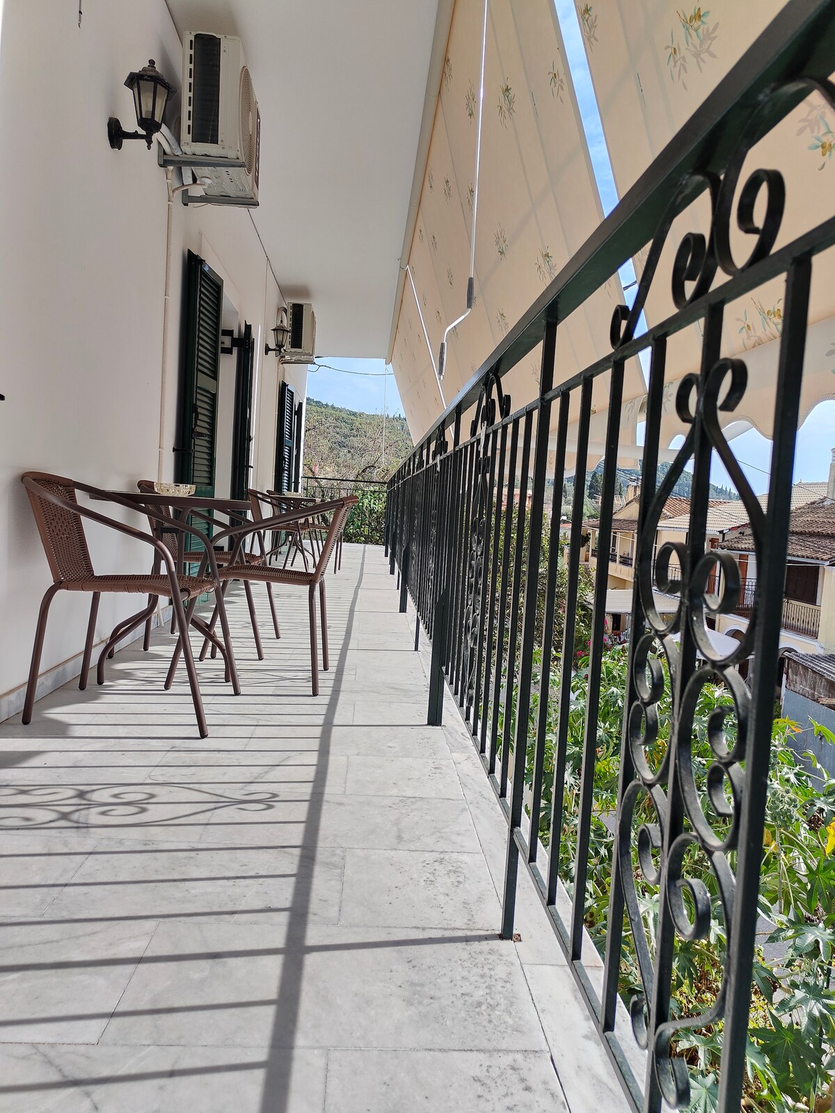 A balcony is featured with a decorative metal railing overlooking the village. Two brown chairs and a small table are positioned to create a relaxing seating area. Sunlight casts shadows on the tiled floor, enhancing the open atmosphere.