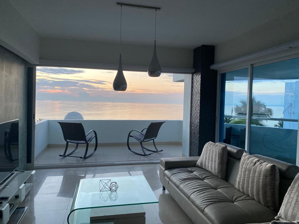 A cozy living area features a modern sectional sofa and a glass coffee table. Two chairs are positioned on the balcony, providing an unobstructed view of the ocean. Large glass doors allow natural light to fill the space, with a stunning sunset visible in the background.