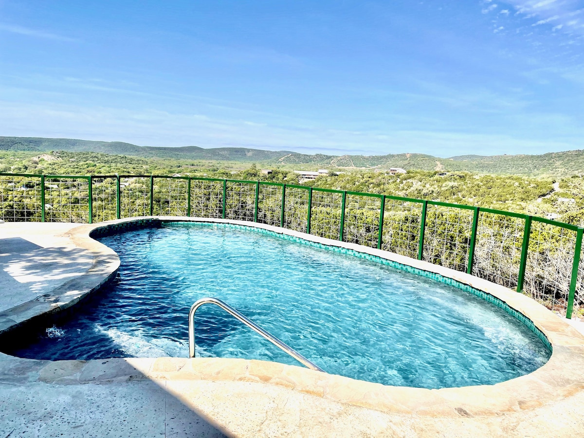 A refreshing pool is set against a scenic backdrop of rolling hills and lush greenery. The pool features a smooth ledge and a single set of stairs leading into the water, creating an inviting space for relaxation.
