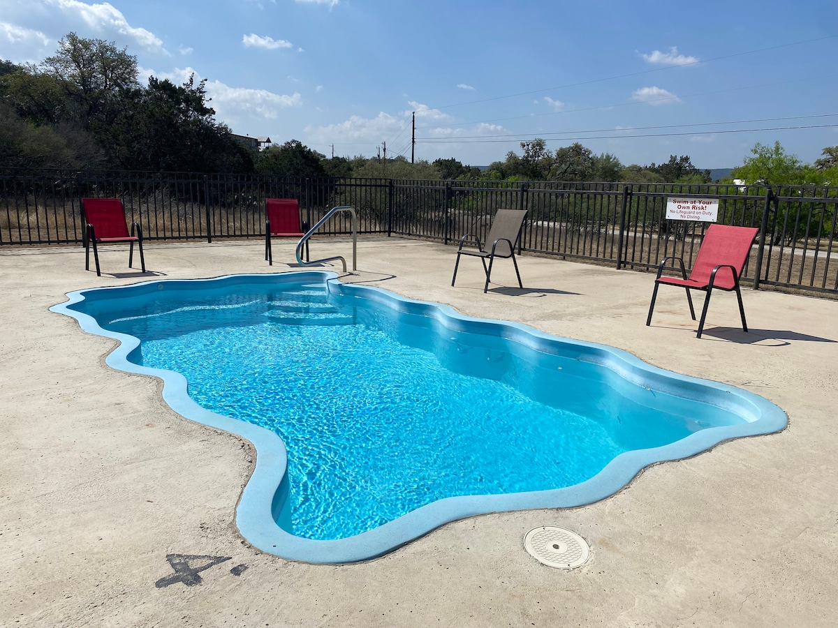 A kidney-shaped pool is surrounded by a concrete deck, featuring five colorful lounge chairs arranged around its perimeter. The crystal-clear water reflects sunlight under a blue sky, while a fence provides privacy and security for outdoor relaxation.