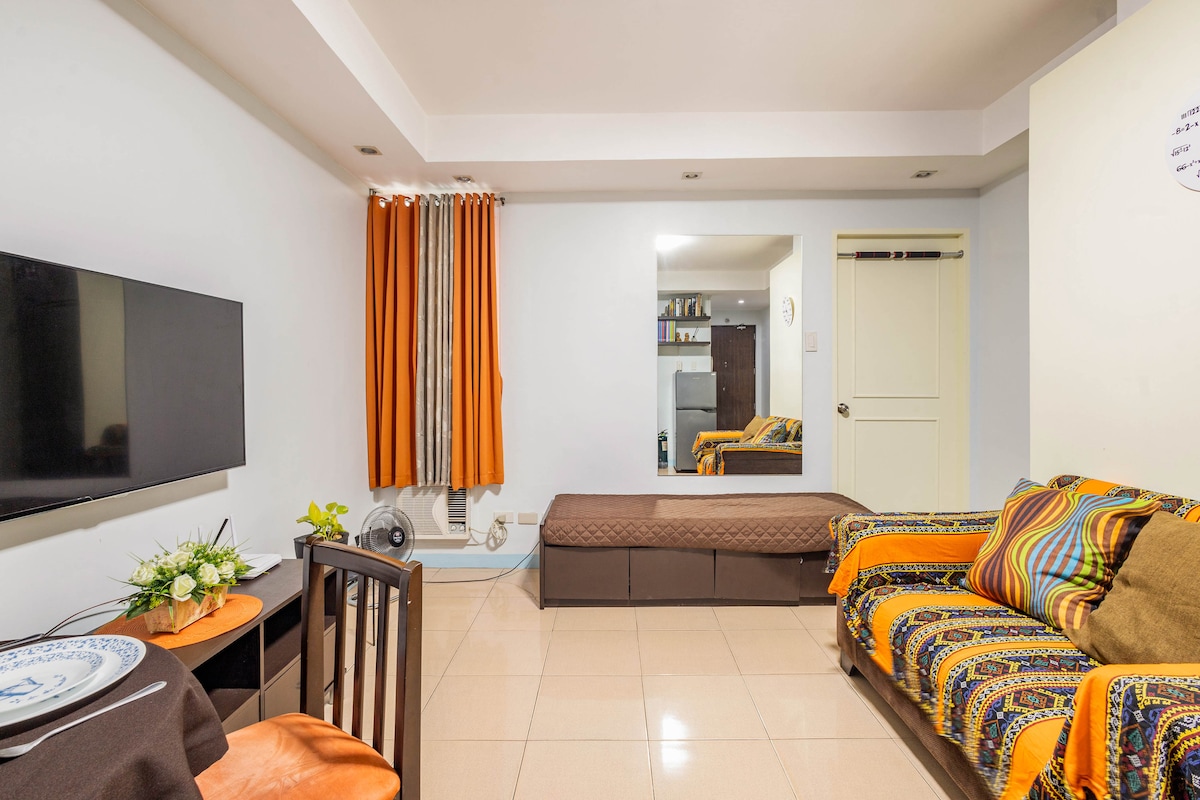 A living area features a large flat-screen television mounted on the wall, with a dark wooden table and a decorative plant nearby. A long cushioned seat is positioned beside an orange-patterned sofa, and curtains provide a pop of color against the neutral walls.
