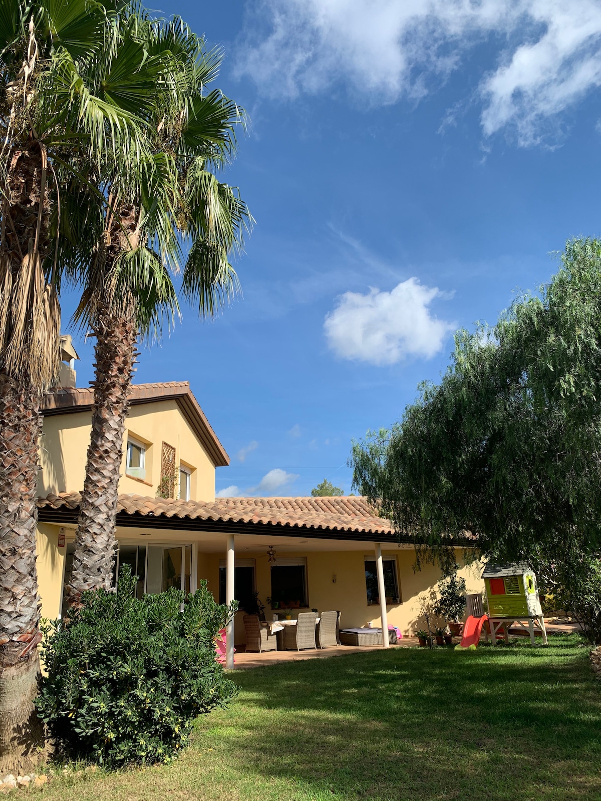 A charming two-story home features a tiled roof and is surrounded by palm trees and lush greenery. A covered patio with comfortable seating is visible, along with a colorful play area in the backyard, all under a clear blue sky.