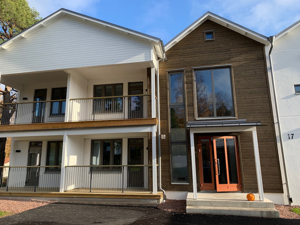 The image shows a modern two-story building featuring a mix of white and wooden exterior finishes. The entrance is framed by large glass windows, and a porch with balcony railings can be seen on the lower level, enhancing the inviting facade.