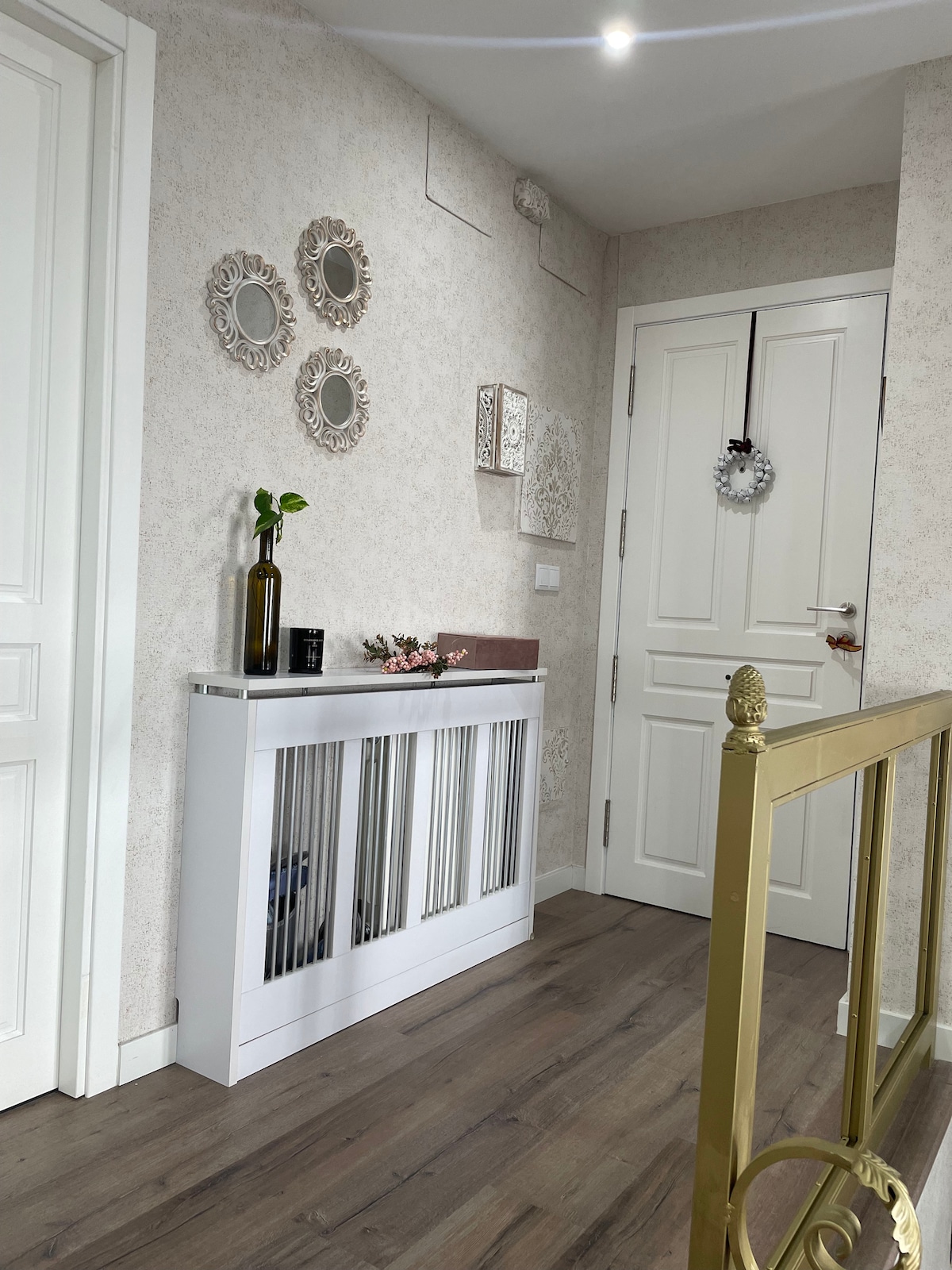A welcoming entryway features a white console table adorned with decorative items, including a vase and floral arrangement. Three ornate mirrors are mounted on the wall, while a well-lit door leads to further spaces. Light wood flooring complements the neutral wall tones.