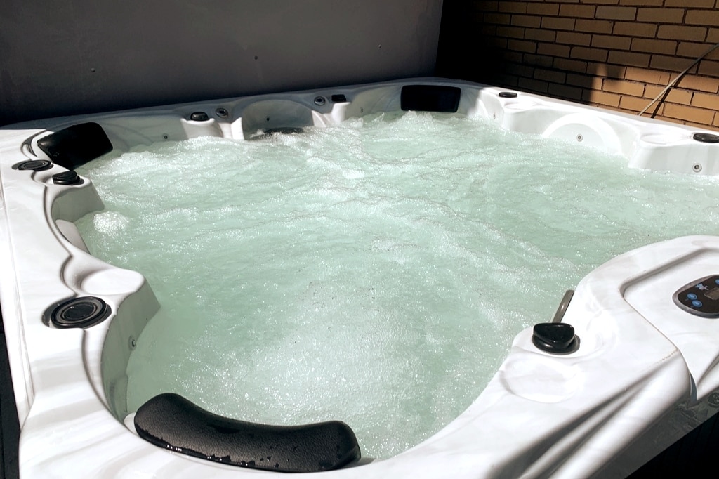A spacious hot tub sits bubbling with clear water, surrounded by smooth edges and built-in seating. Jets are visible, ready for relaxation. The background features brick walls, adding a touch of warmth to the soothing setting.