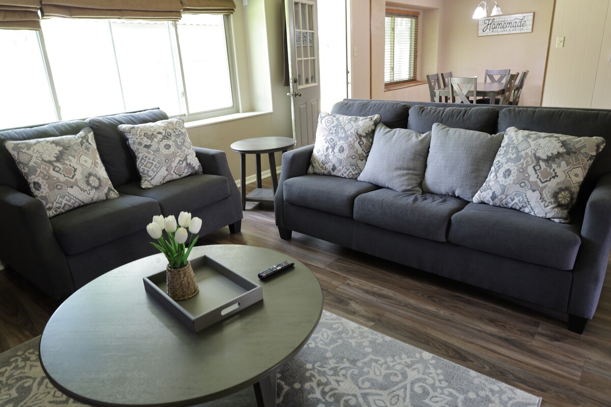 A spacious living area features two comfortable sofas adorned with patterned cushions. A round coffee table sits in front, with a small tray and a decorative vase holding tulips. Natural light streams through large windows, creating an inviting atmosphere.