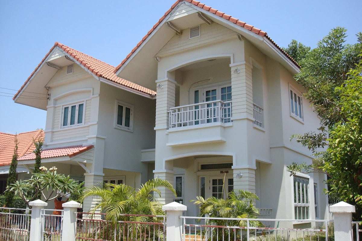 A two-story house features a combination of white and muted pastel siding, with a red-tiled roof. A spacious balcony supports decorative railings, and palm trees are visible in the well-maintained garden. The front porch extends a welcoming feel, framed by green foliage.