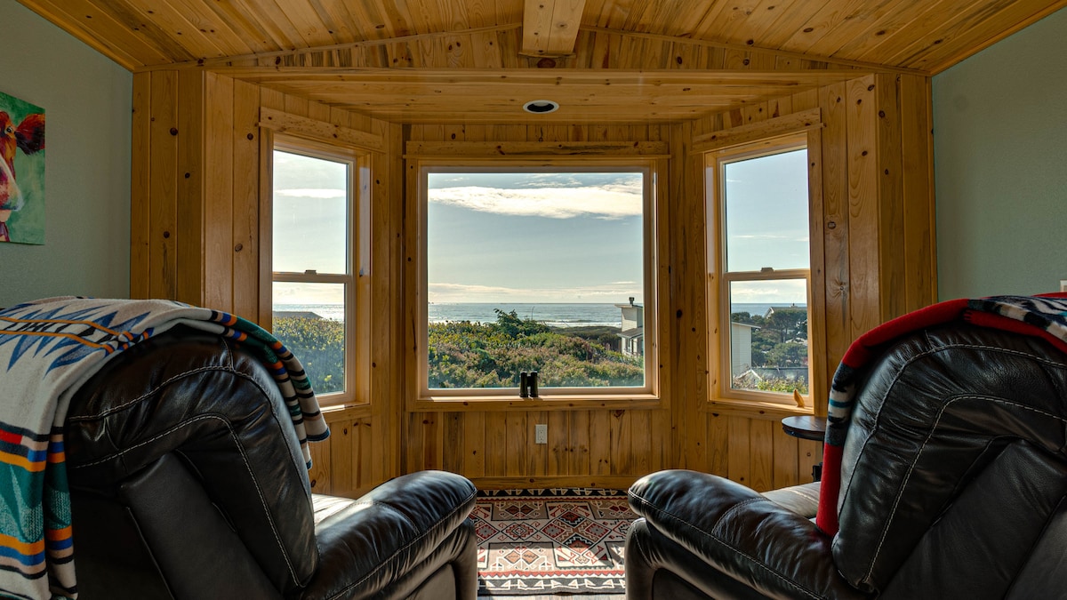 A cozy seating area features two overstuffed recliners positioned to face a large window. Natural light fills the space, revealing views of the ocean and coastal landscape. The wooden walls and ceiling add warmth, while a patterned rug complements the rustic charm.