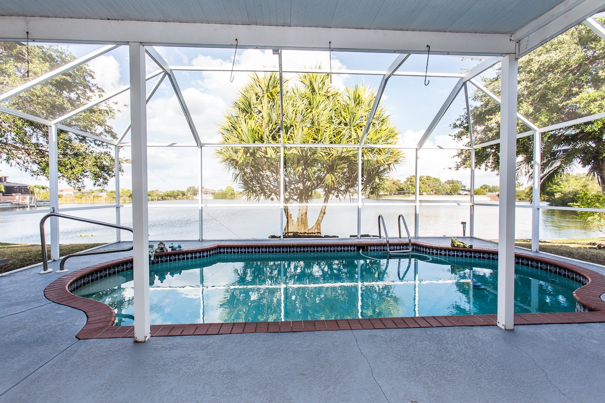 A screened pool area features a sparkling pool, surrounded by concrete flooring and framed by palm trees. The tranquil lake view is visible through the mesh enclosure, offering a serene backdrop for relaxation. Sunlight filters through, creating a warm ambiance.