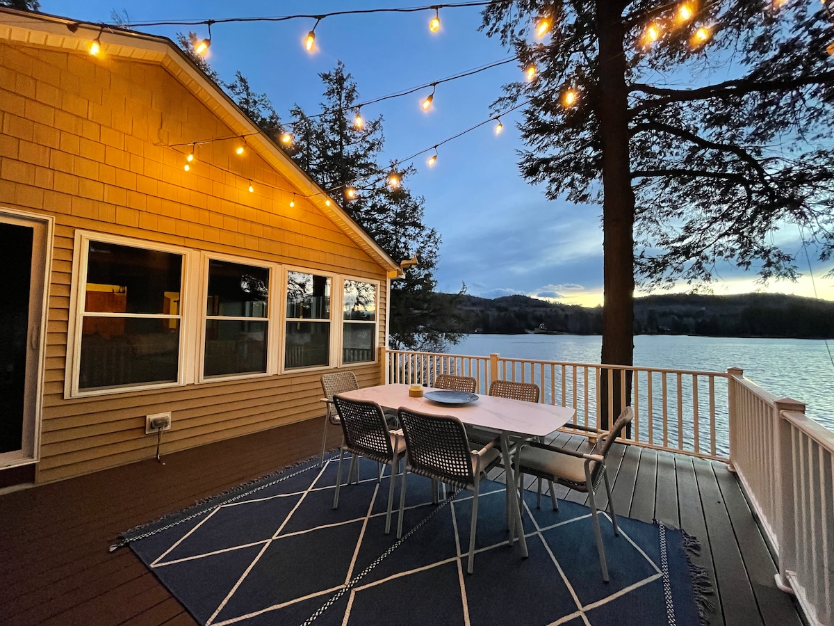 A spacious deck features a dining table surrounded by metal chairs, illuminated by string lights overhead. The tranquil lake reflects the evening sky, while large windows provide a view of the interior. A decorative area rug enhances the outdoor space.