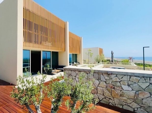 A modern property is shown featuring horizontal wooden slats on the upper exterior, creating a distinctive design. Large glass windows provide views of the surrounding landscape, which includes greenery and a stone wall. The ocean is visible in the background under a clear blue sky.