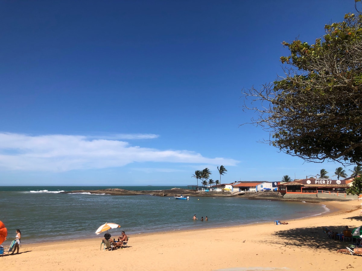 A sandy beach is observed, with calm waters gently lapping at the shore. Sun umbrellas provide shade for visitors relaxing on the sand. A few people can be seen in the water, while palm trees sway gently in the breeze against a clear blue sky.