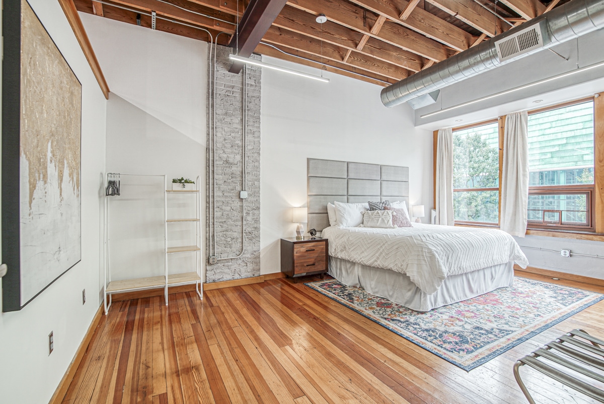 A spacious bedroom features a large bed dressed in white linens, complemented by soft bedside lamps on wooden nightstands. Natural light enters through wide windows adorned with light curtains, illuminating polished wooden floors. A wall-mounted shelf stands nearby, and exposed wooden beams add to the room's character.
