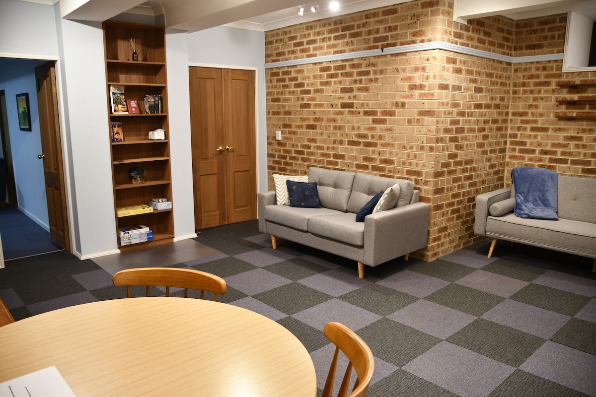 A spacious living area features a grey sofa and an additional light grey couch, positioned against a brick wall. A circular dining table with wooden chairs is visible in the foreground. Natural light is supplied through the doorway and supplemental lighting enhances the room's comfort.