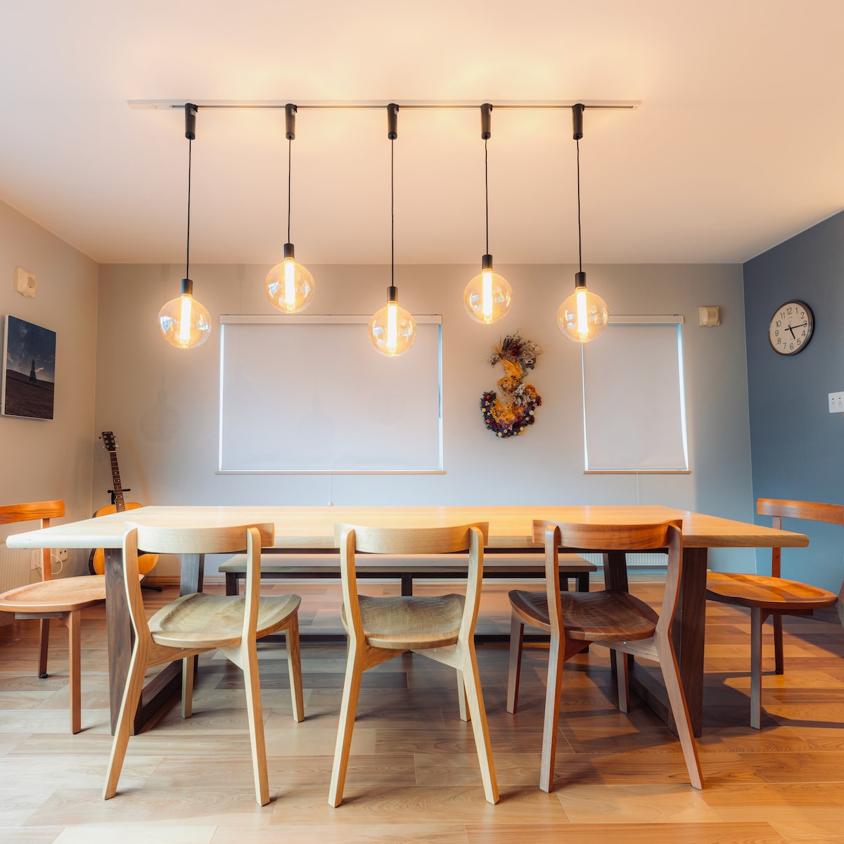 A dining area showcases a long wooden table surrounded by six wooden chairs. Above, pendant lights provide illumination, while large windows allow natural light to enter. A clock and decor adorn the walls, contributing to the functional and inviting space.