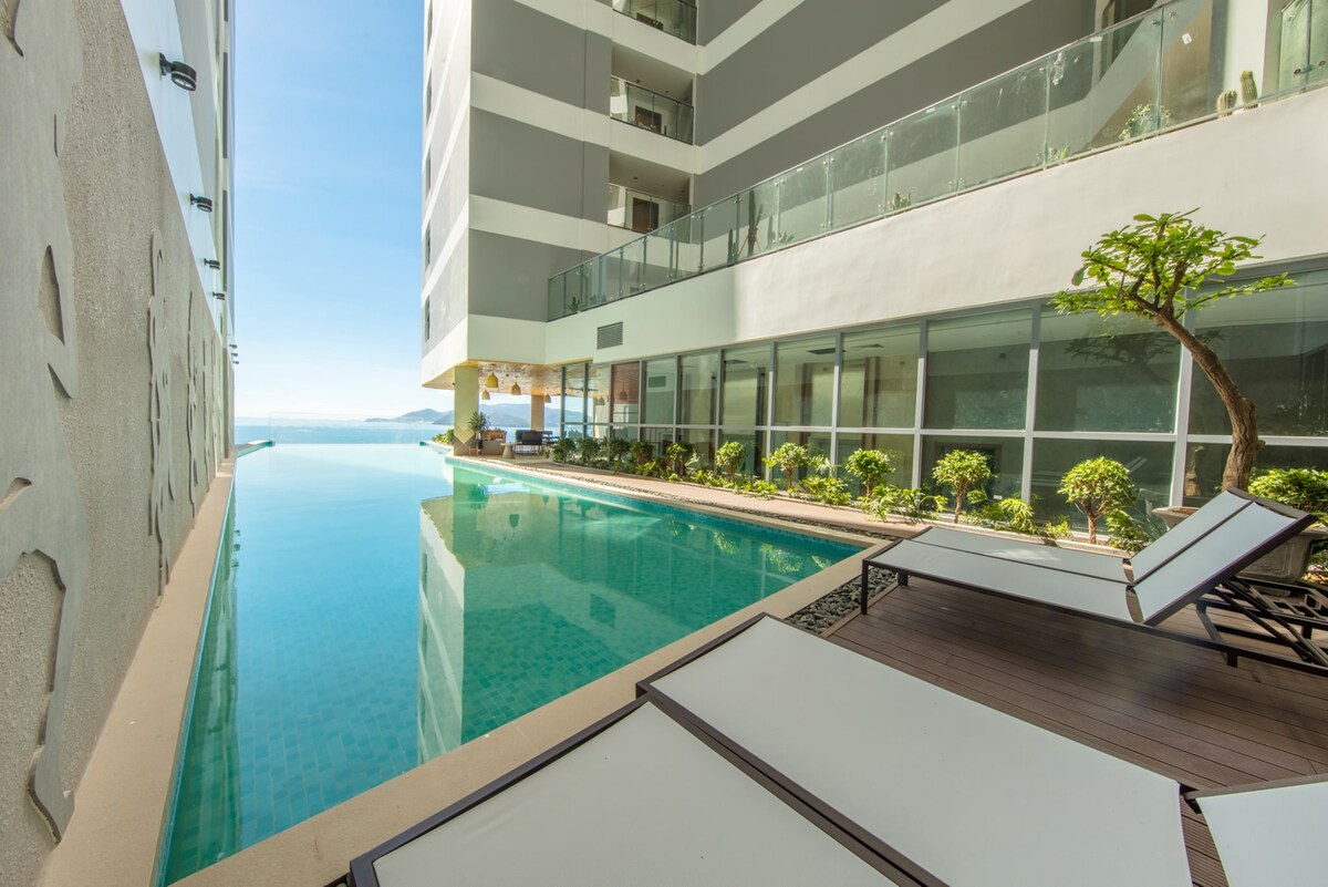 A serene pool extends alongside the building, surrounded by well-maintained greenery. Sun loungers are positioned near the water's edge, inviting relaxation. The calm surface of the pool reflects the architecture and the clear sky above, providing a tranquil outdoor space.