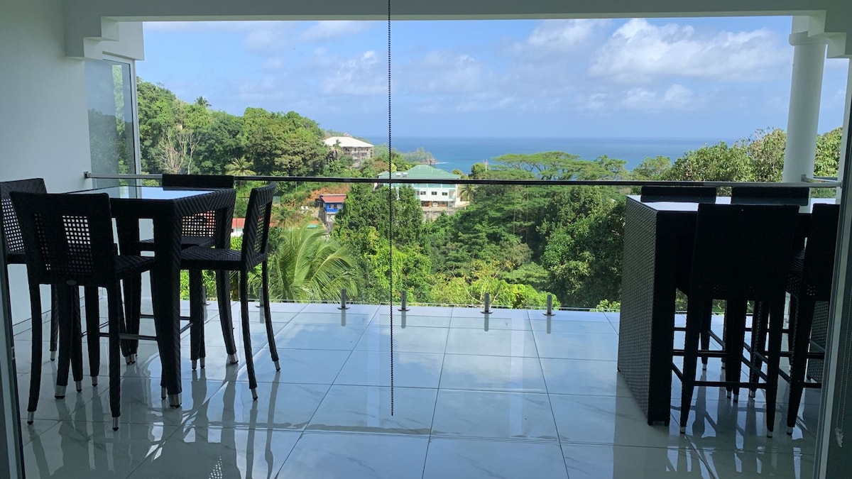 A spacious balcony displays several high-top tables and chairs, offering an unobstructed view of lush greenery and the ocean in the distance. Large glass panels enhance the openness of the area, allowing natural light to fill the space.