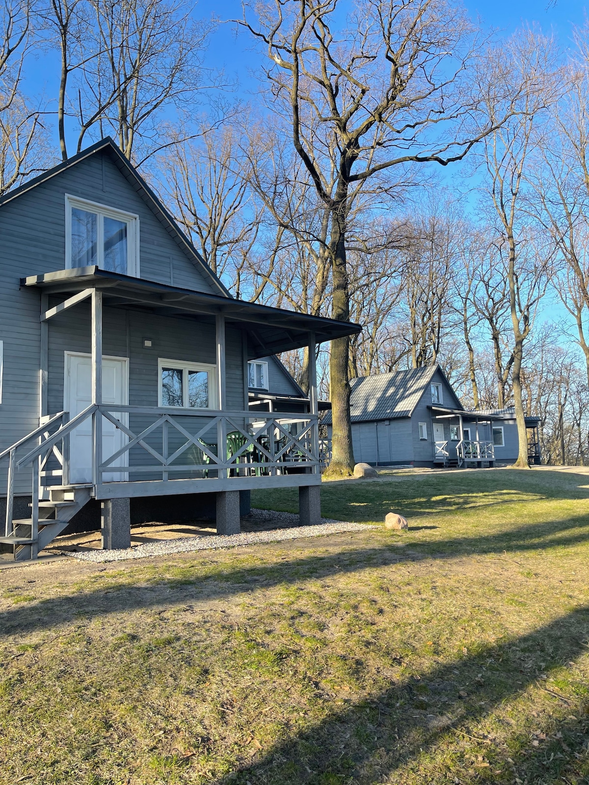 Two cabins with gray exteriors are positioned on a grassy hillside. Each cabin features a front porch with railings and large windows. Sparse trees surround the area, and a clear blue sky is visible above.