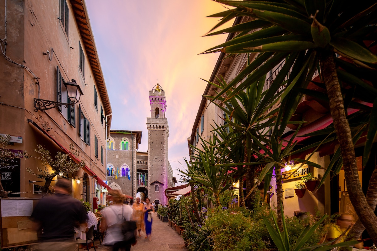 A charming street is lined with vibrant green plants and restaurants, showcasing the historical architecture of a bell tower in the distance. The sky is painted with soft hues of pink and orange, reflecting the onset of dusk.