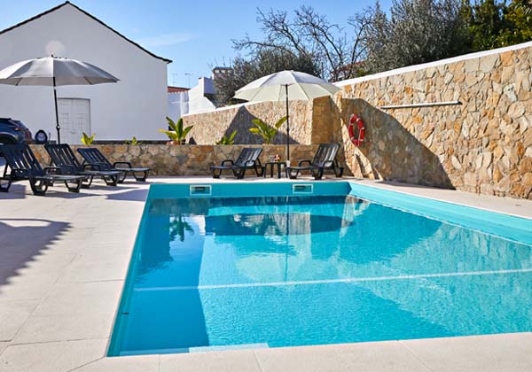 A clear swimming pool reflects the sunlight, bordered by a spacious pool deck. Several lounge chairs are arranged beside the water, accompanied by large umbrellas providing shade. A stone wall adds texture to the area, enhancing outdoor relaxation options.