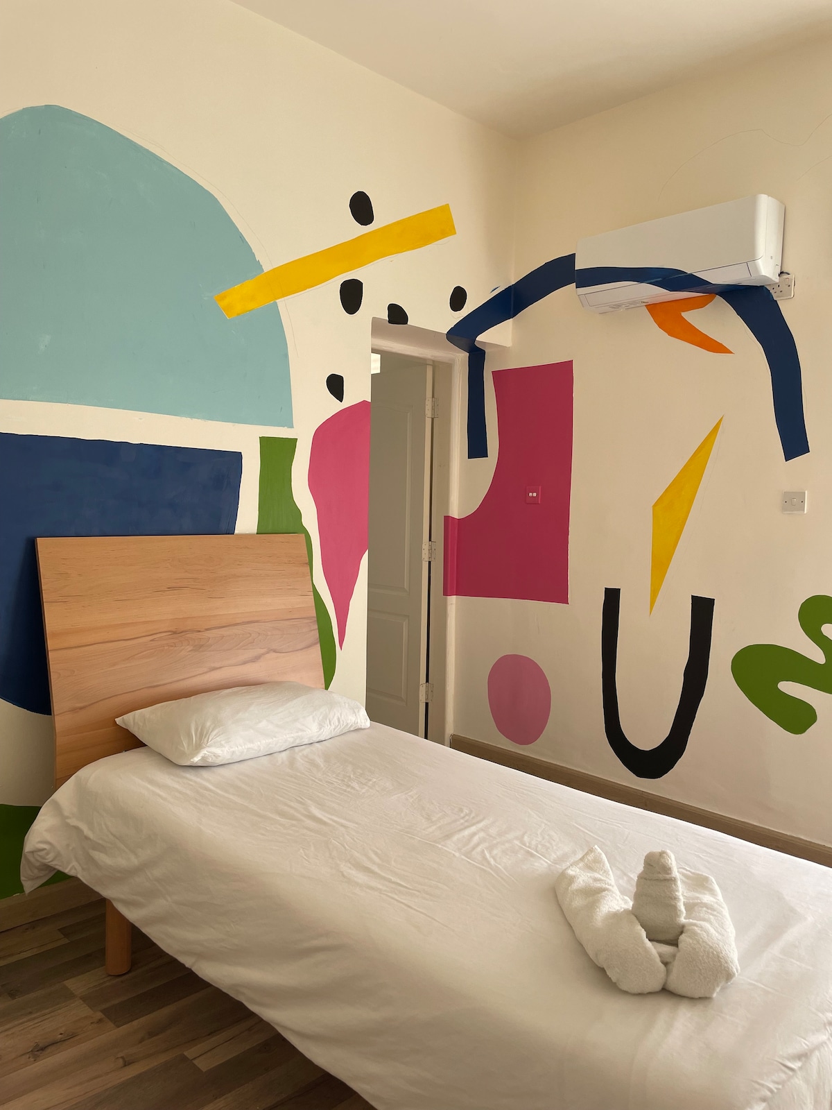 A vibrant sleeping area features a single bed with a light-colored blanket. The wall behind the bed is adorned with colorful abstract shapes, creating an engaging visual impact. A door leads to another room, while an air conditioning unit is positioned above.