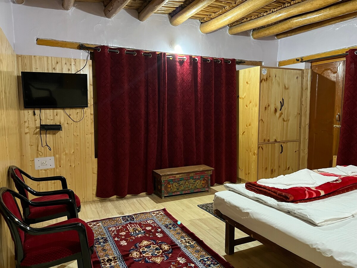 A cozy room features wooden accents and traditional decor, including a vibrant rug. A spacious bed with fresh linens is present, along with two black chairs positioned nearby. A television is mounted on the wall, and rich red curtains provide privacy.