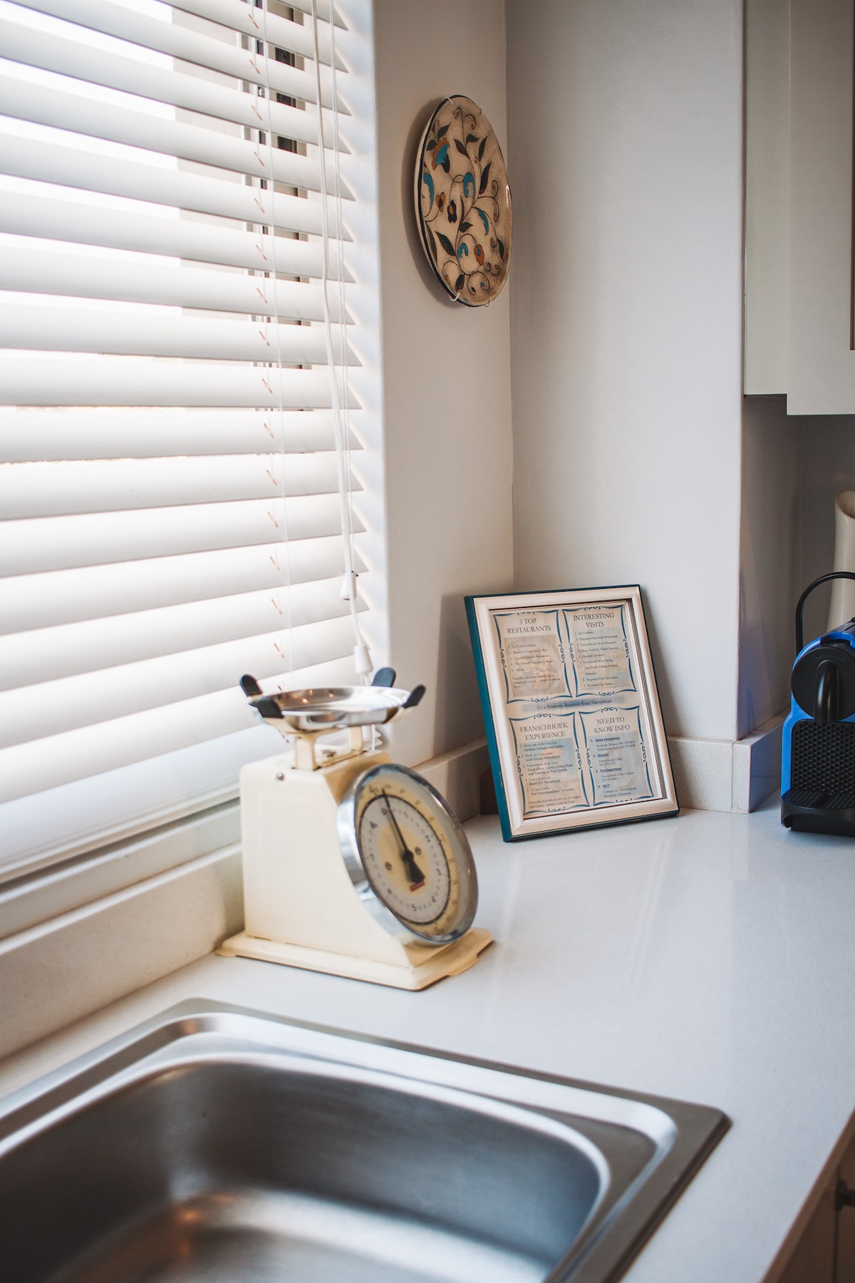 A clean kitchen area features a stainless steel sink and a classic scale on the countertop. Sunlight filters through blinds, creating a warm ambience. A framed menu or information sheet is displayed on the wall, adding a personal touch.