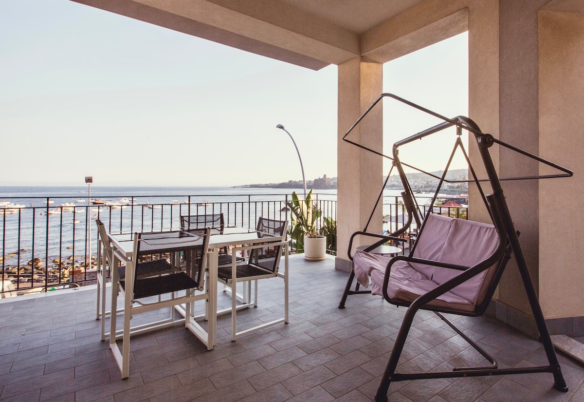 A spacious terrace offers seating arrangements including a swing chair and a dining table with chairs. The view overlooks the calm sea and nearby coastline, providing an inviting space for relaxation. Sunlight gently fills the area, enhancing the overall ambiance.
