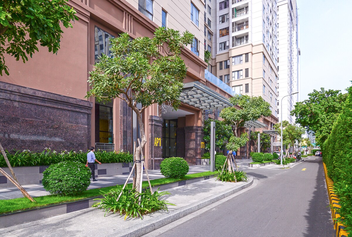 A well-maintained entrance to a modern building is surrounded by neatly trimmed shrubs and small trees. A wide walkway leads to the main entrance, which is sheltered by a glass canopy. The street is flanked by decorative landscaping, enhancing the urban environment.