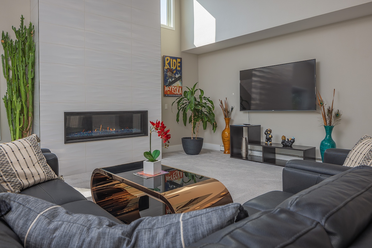 A modern living area features a sleek gas fireplace set within a light-colored wall. Comfortable black sofas are arranged around a reflective coffee table, complemented by decorative plants and vases. Natural light illuminates the space through high windows.