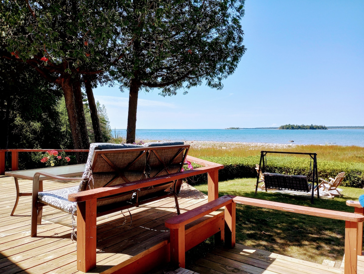 A spacious deck overlooks Lake Huron, featuring comfortable outdoor seating with cushions and a relaxing swing. Lush greenery frames the view of the water, dotted with islands in the distance. The inviting space offers an ideal setting for enjoying the outdoors.