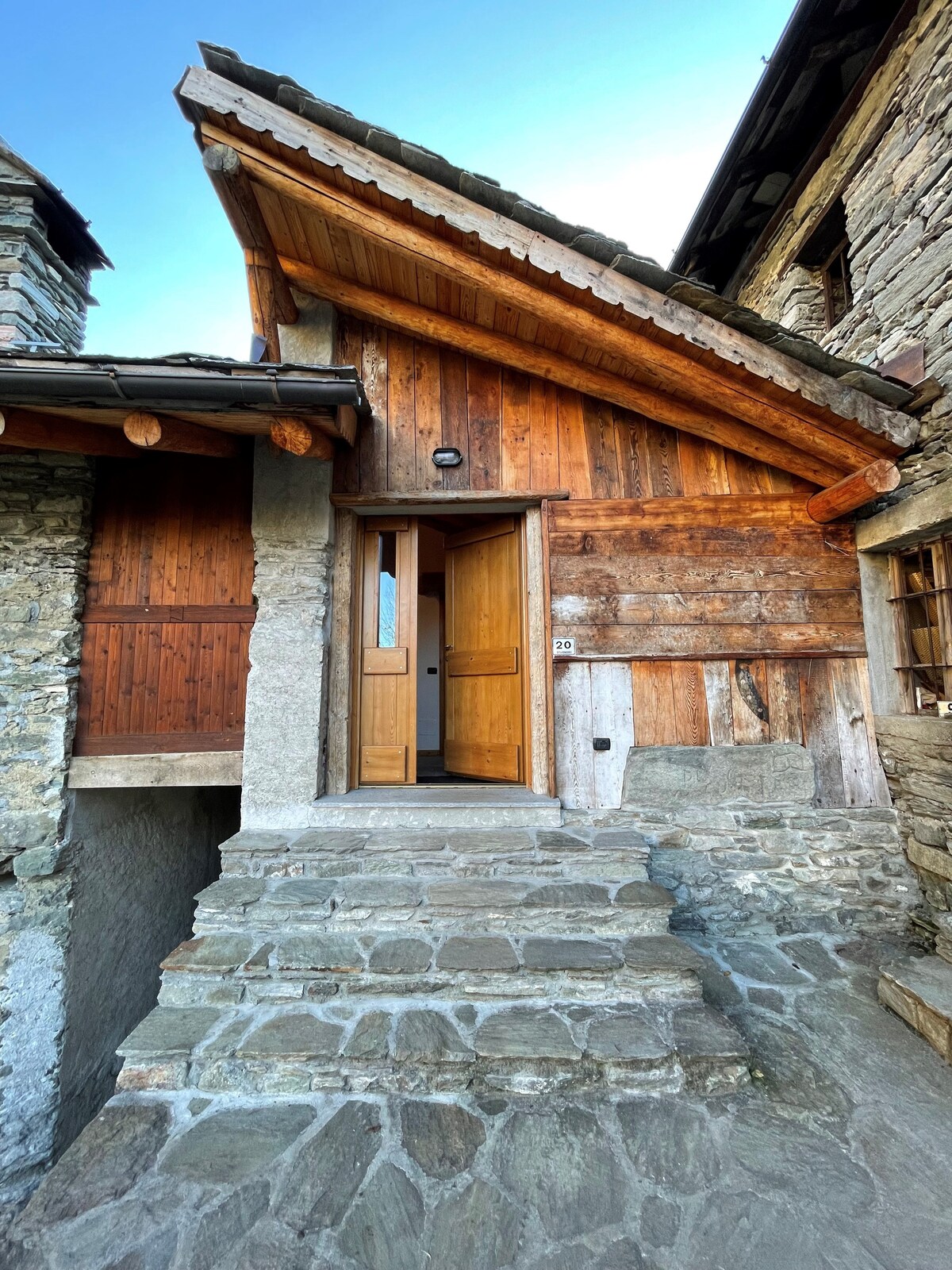 The entrance of a rustic stone cottage features a wooden door set within a sloped roof structure. Stone steps lead to the entrance, bordered by a combination of rough stone and wooden elements, reflecting traditional architectural style. Natural light brightens the entryway.