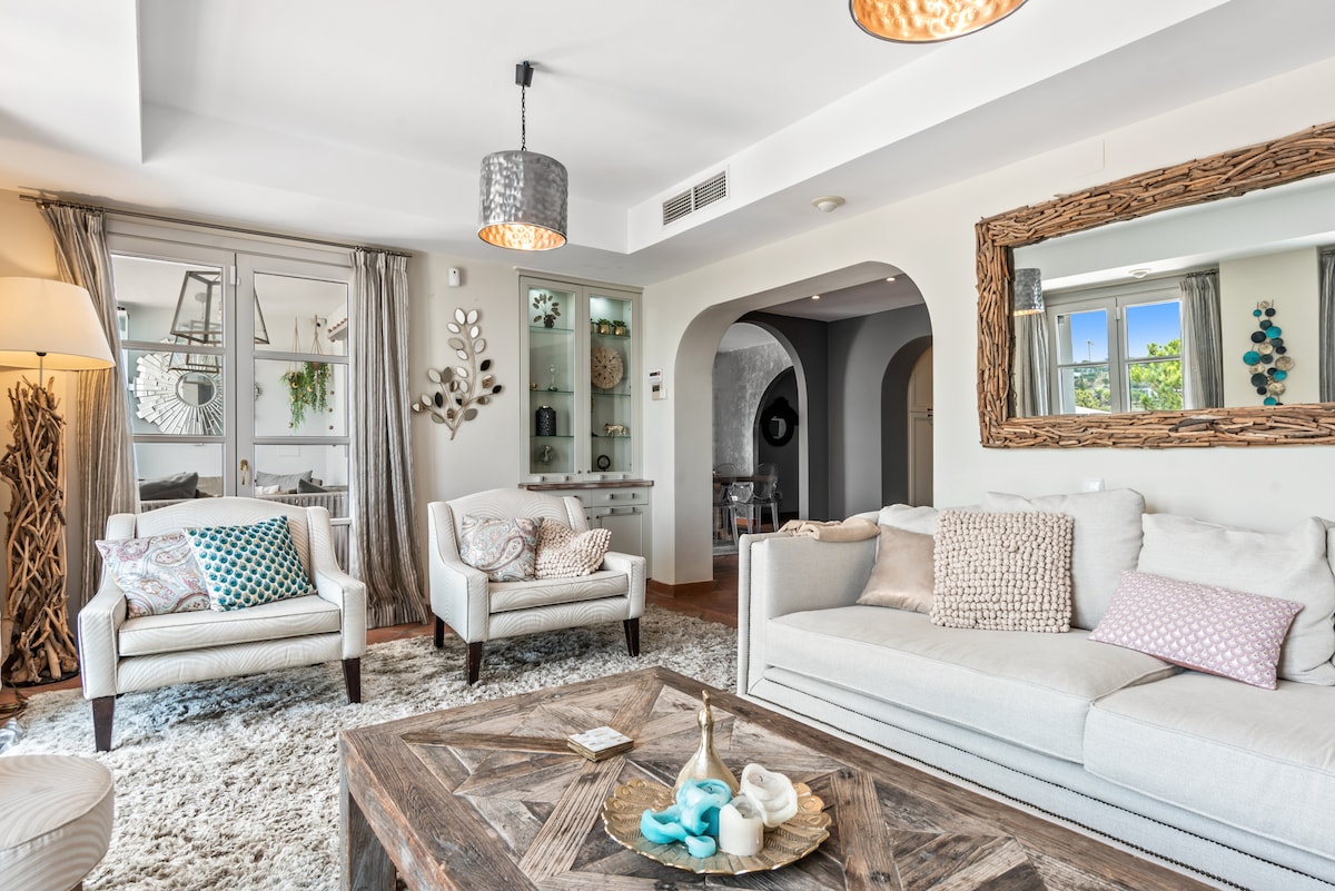 A light-filled living room features a spacious white sofa adorned with various textured cushions. Two armchairs with decorative pillows are positioned across from a wooden coffee table. Natural light enters through large windows decorated with sheer curtains, creating a warm and inviting space.