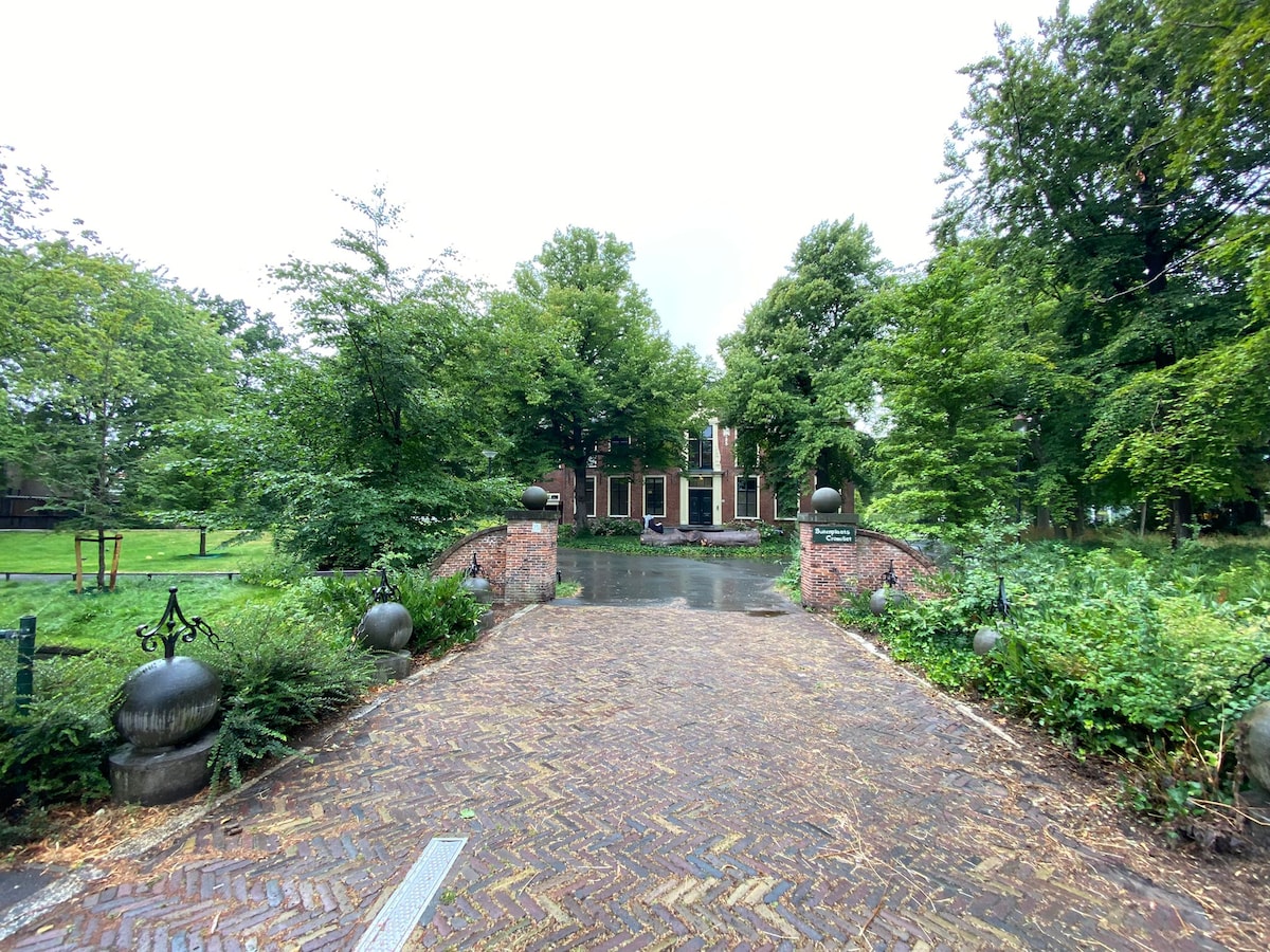 A cobblestone pathway leads to a welcoming brick house surrounded by lush greenery. Tall trees and well-maintained shrubs frame the entrance, creating a serene atmosphere. A gentle slope guides visitors towards the main building, which is partially visible through the foliage.