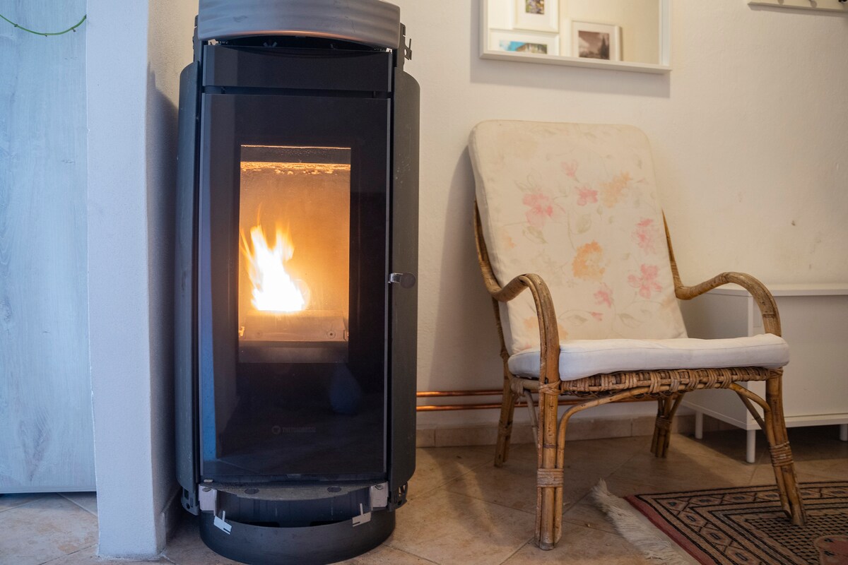 A modern pellet stove is positioned next to a comfortable rattan chair. The stove emits a warm glow from its glass front, creating an inviting focal point for the cozy living area.