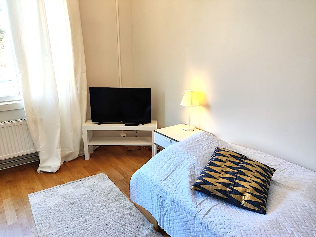 A well-lit sleeping area features a single bed covered with a white quilt and a decorative pillow. A small rug lies beneath, and a modern television is positioned on a white entertainment unit. Soft curtains allow natural light to brighten the space.