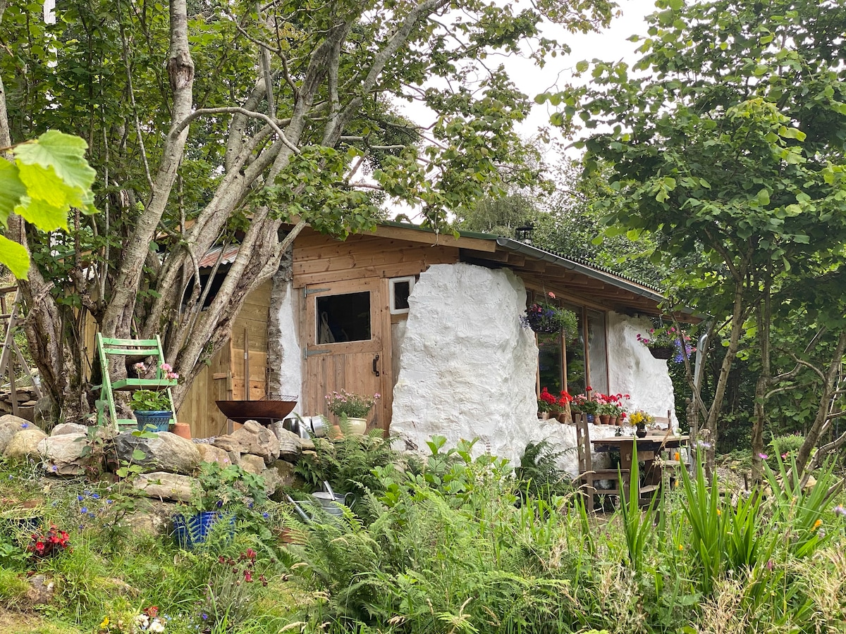 The bothy’s exterior is surrounded by lush greenery, featuring a mix of stone and wooden walls. Flower boxes adorn the windows, adding vibrant splashes of color. A simple table sits outside, inviting peaceful moments amidst natural beauty.