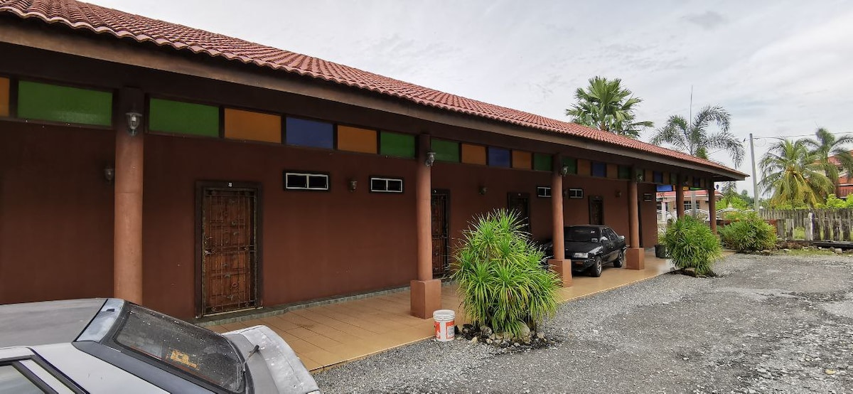 The exterior of the building features a long, rectangular facade with contemporary colors accenting the upper section. Several wooden doors are framed by tall, round columns, and lush green plants are arranged in front, providing a natural touch. A parking area with vehicles is visible in the foreground.