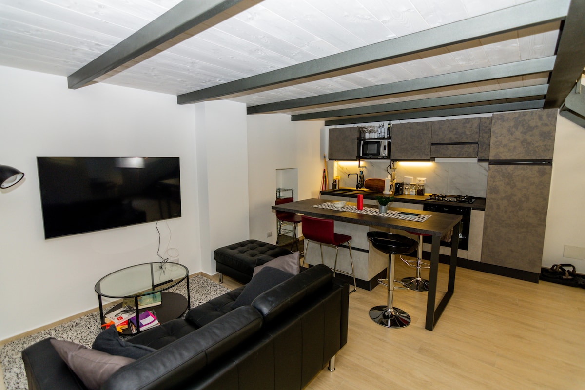 An open space layout is shown, featuring a modern kitchen with sleek cabinetry and integrated appliances. A comfortable black leather sofa faces a wall-mounted television. A glass coffee table rests on a textured rug, enhancing the clean, contemporary design of the living area.