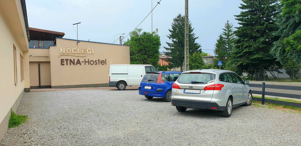 A gravel parking area is presented, accommodating multiple vehicles. The exterior of the ETNA Hostel, featuring a prominent sign, is visible along with trees in the background and utility poles. A clear sky is seen above, adding a sense of openness.
