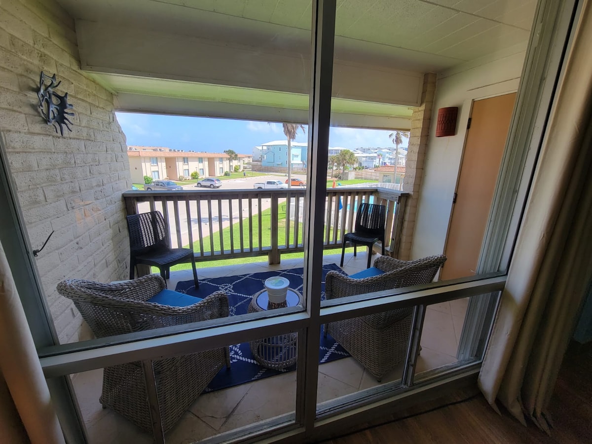 A balcony space features two woven chairs positioned around a small table. A decorative rug is placed beneath, and a view of the surrounding area is visible through the railing. Natural light enhances the inviting atmosphere.