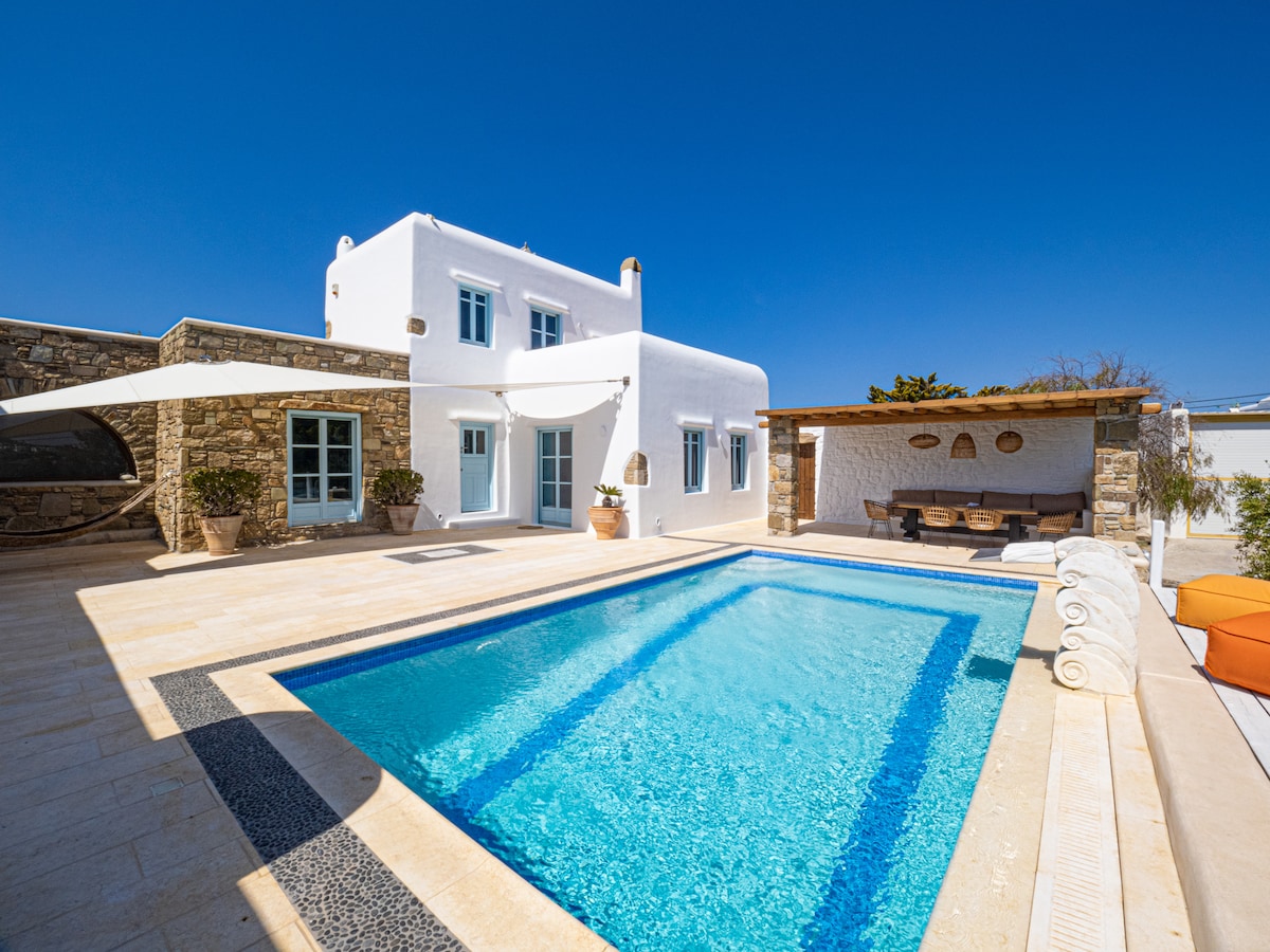 A modern villa features a spacious swimming pool surrounded by a sun terrace. The exterior showcases a whitewashed structure with large windows and a shaded outdoor seating area nearby. Clear blue skies enhance the atmosphere, while a stone wall adds texture to the setting.